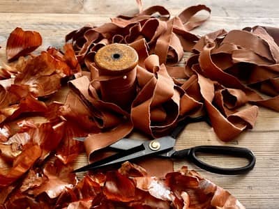 A vintage wooden thread spool wrapped in naturally dyed terracotta ribbon, surrounded by dried onion skins and a pair of dressmaking scissors on a pale wooden surface.