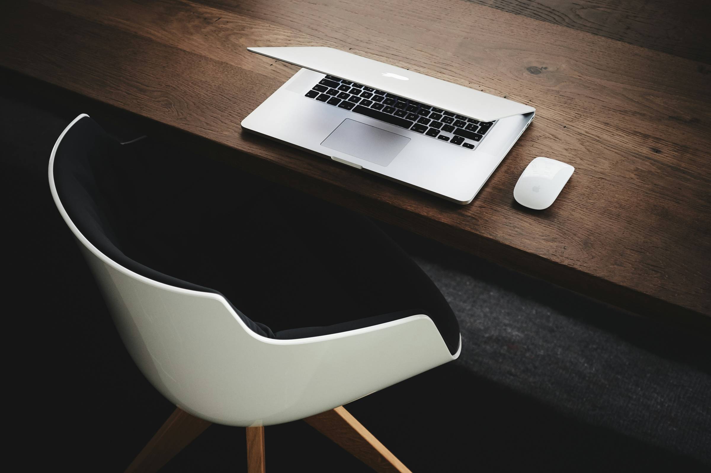 An empty white chair sits at a desk. A laptop and mouse are visible from the view looking down to the desk.