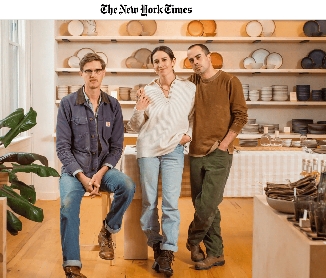 Three people stand - all wearing jeans and casual shirts - in front of a wall of East Fork pottery.