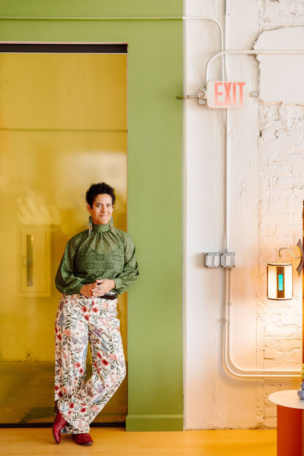 Naomi stands in front of a yellow glass wall and a green wall, with a painted white brick exposed wall to her left. She is wearing a green top and floral print pants.