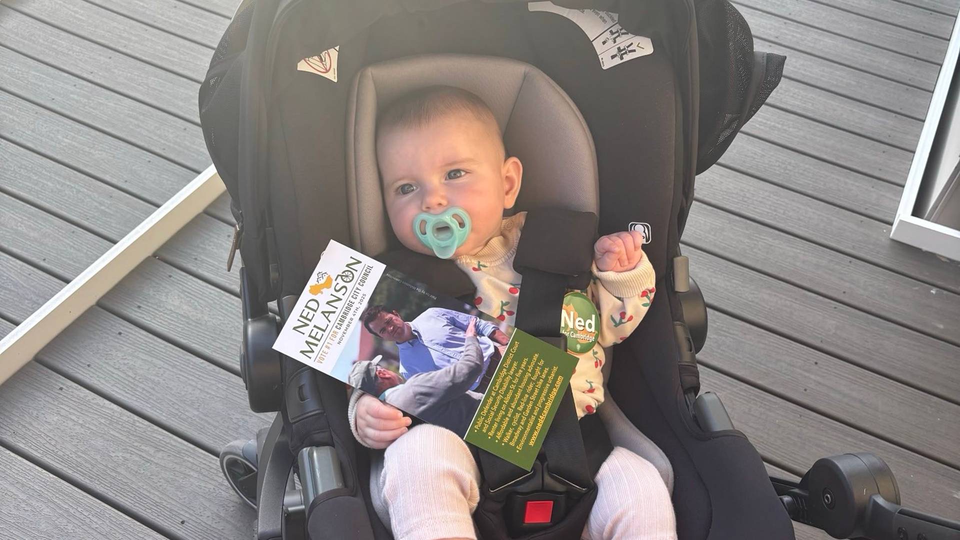 A baby in a carrier wearing a Ned pin