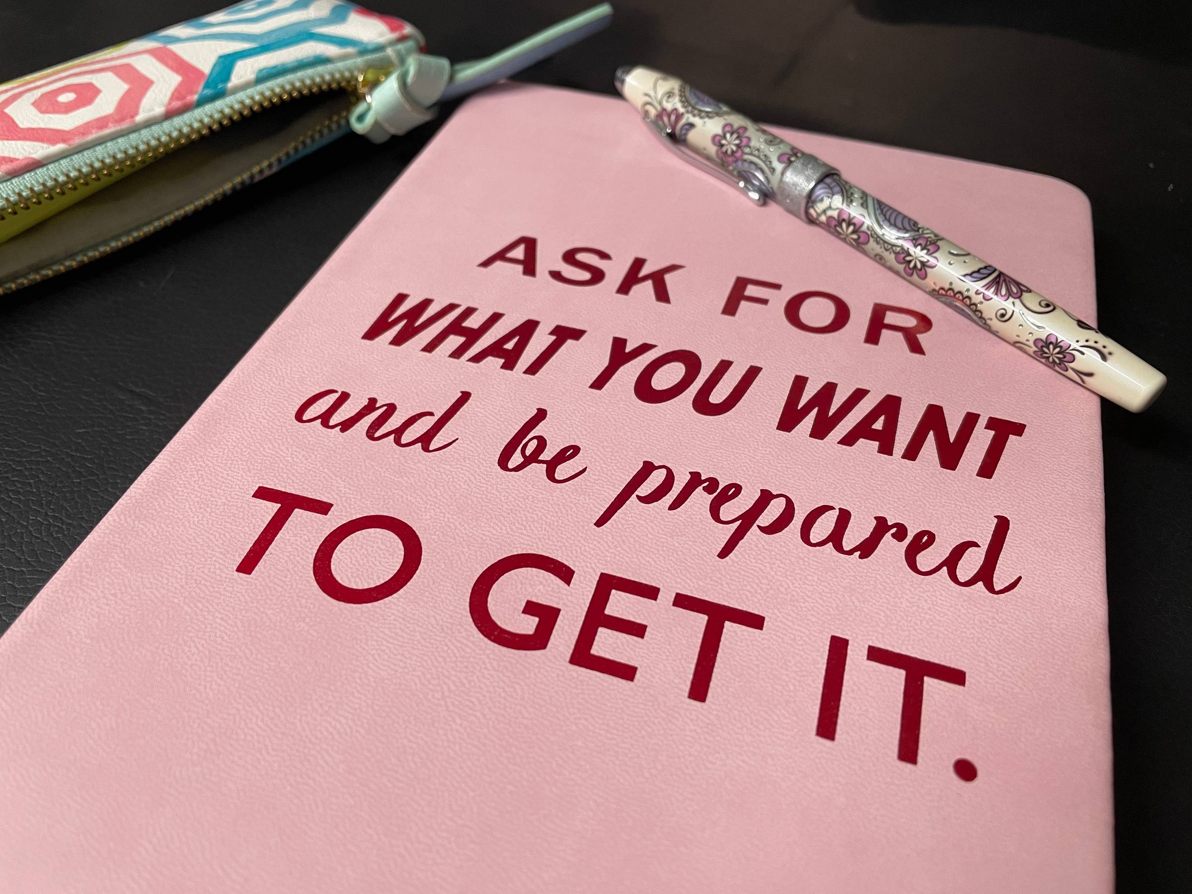 Pink notebook that says:"Ask for what you want and be prepared to get it." with a colourful pen on top and pencil case to the side.