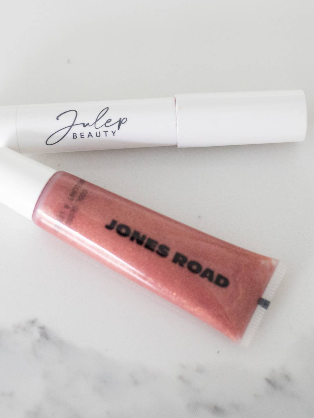 Julep Beauty tinted balm in white tube and Jones Road lip gloss in shimmery pink tube on a white background.