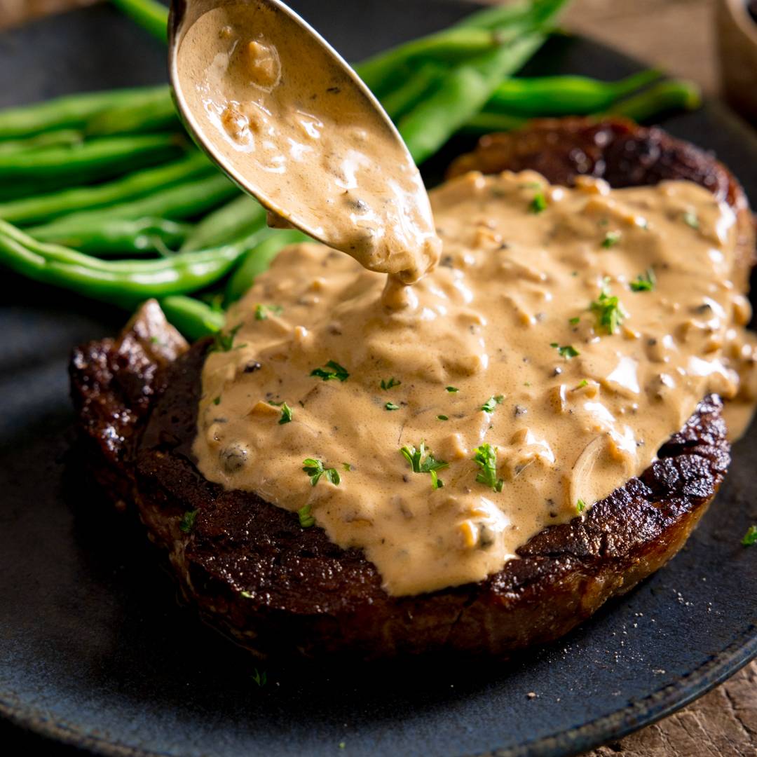Peppercorn sauce being poured over a steak.