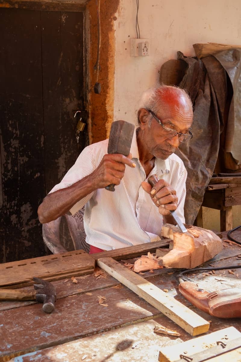 Elderly man carves wood with hammer and chisel.