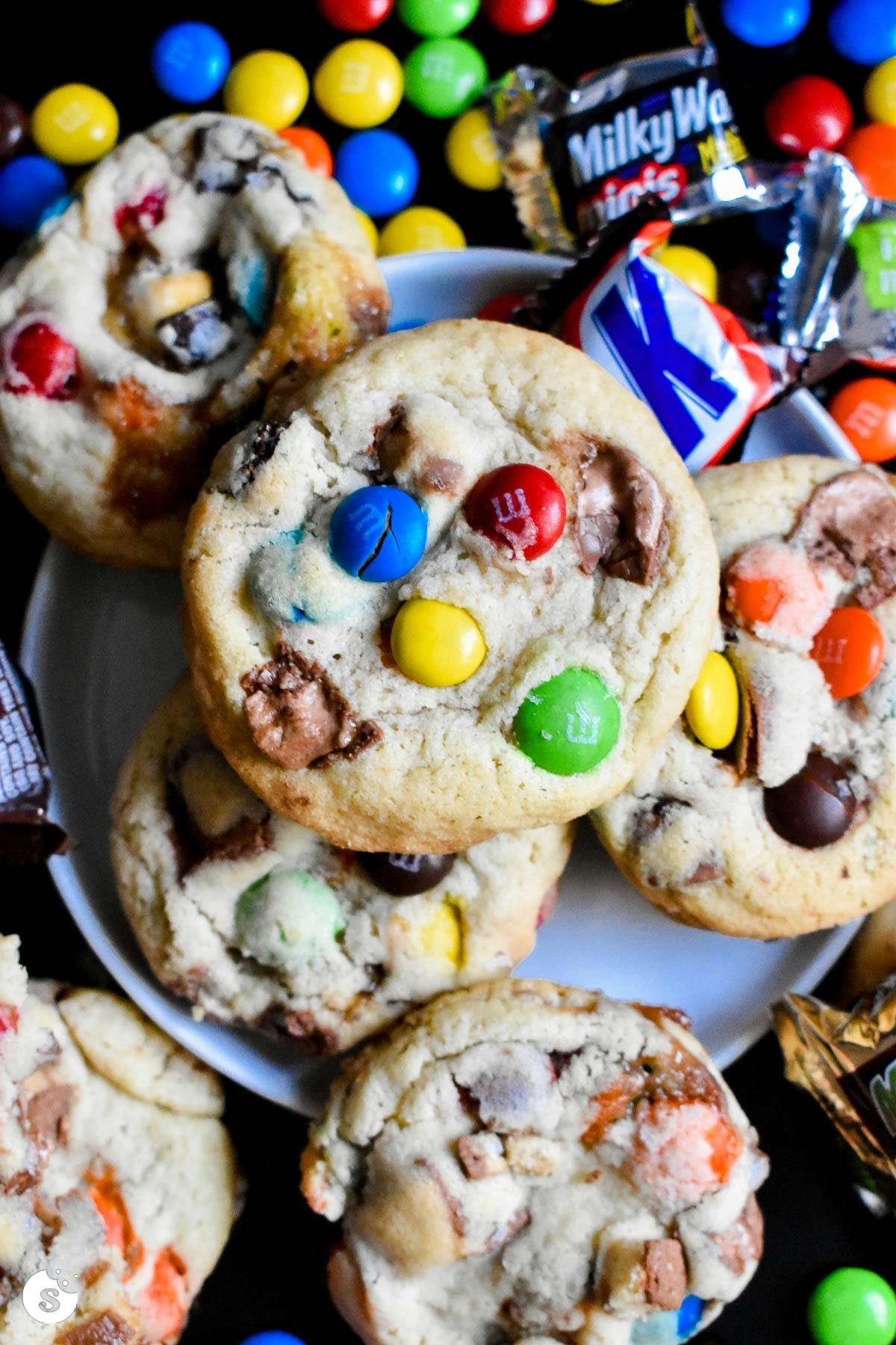 Cookies stuffed with chocolate and M&Ms piled on a black surface surrounded by candy bars and M&Ms