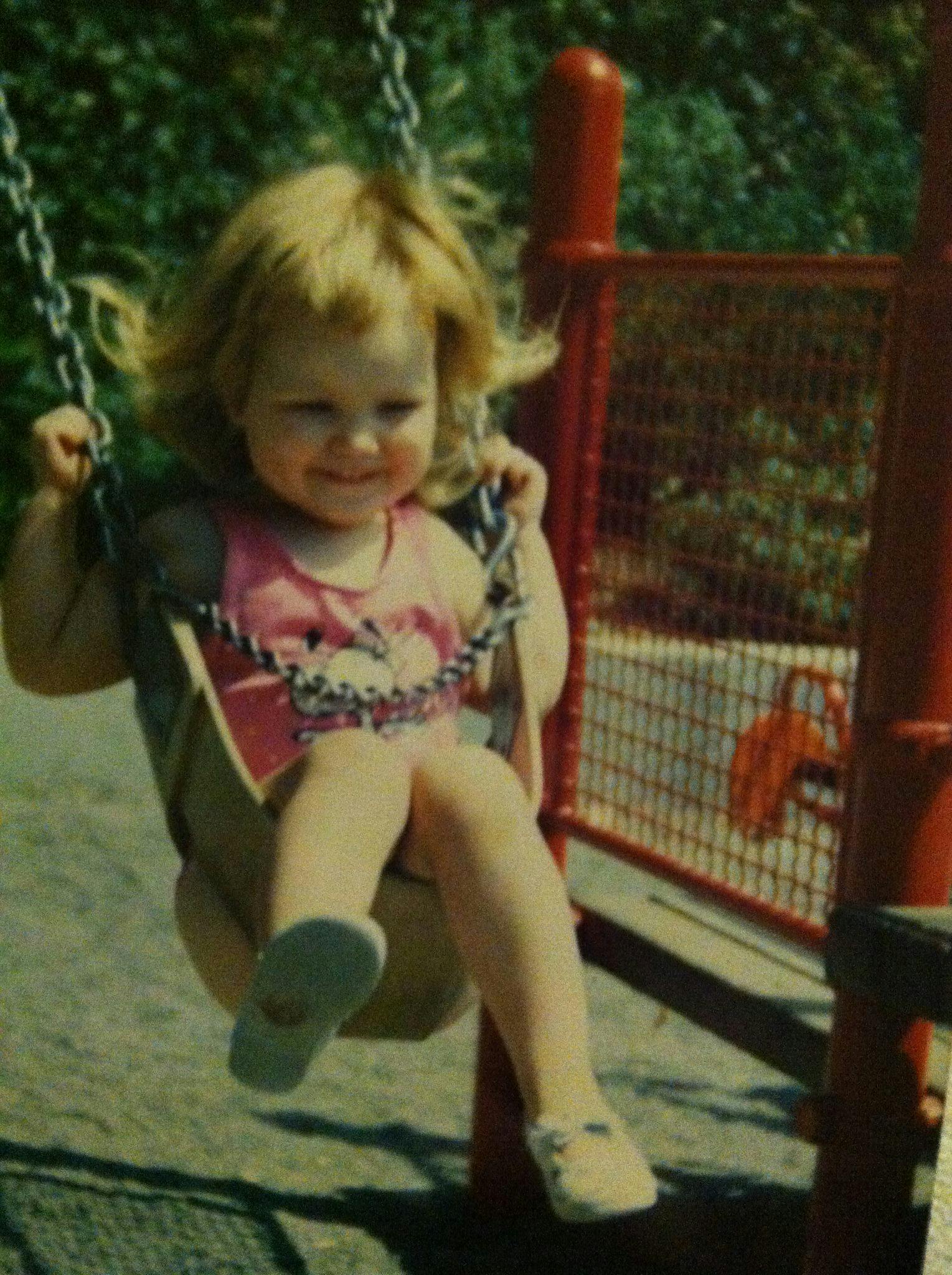 Lucy at two years old with blond hair and pink Snoopy tank top, riding in a swing and smiling