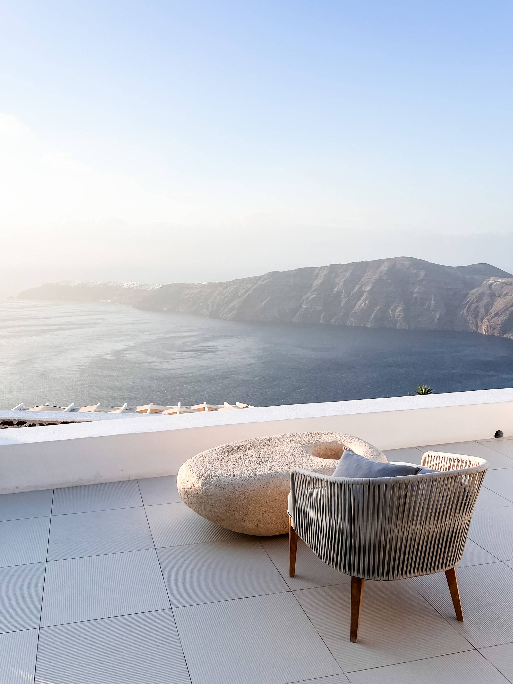 A serene outdoor setting featuring a modern wicker chair with a cushion, positioned on a tiled terrace. Next to the chair is a round, textured stone table. The terrace overlooks a breathtaking view of the sea, leading to rugged, distant hills under a clear blue sky. The lighting suggests a tranquil and sunlit atmosphere, possibly during early morning or late afternoon.