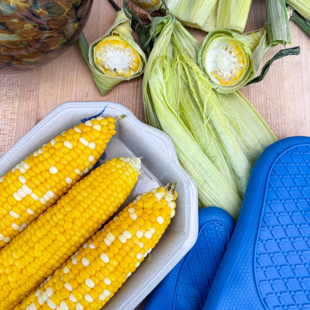 beautiful silk free sweet corn cobs on plate with blue oven mitts and husk with silk in it