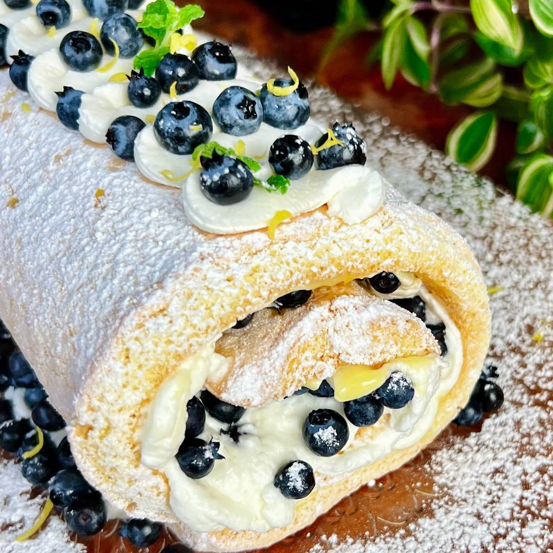 blueberry Swiss roll with end showing whipped cream, lemon curd and garnished with fresh blueberries, mint and powdered sugar