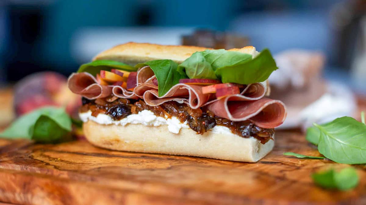 A gourmet open-faced prosciutto sandwich on a rustic ciabatta roll, layered with goat cheese, prune and shallot jam, fresh peach slices, and basil leaves, served on a wooden cutting board.