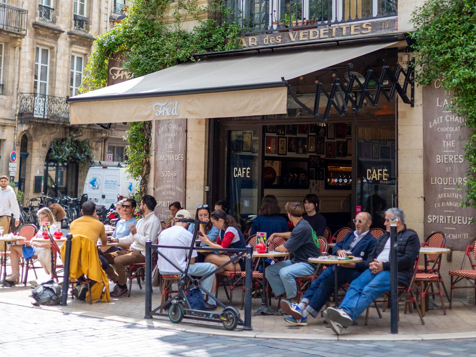 A lively outdoor terrace at Bar des Vedettes café in Bordeaux, France, with groups of people dining and drinking beneath a cream awning, framed by classic Haussmann-style limestone architecture and lush greenery.