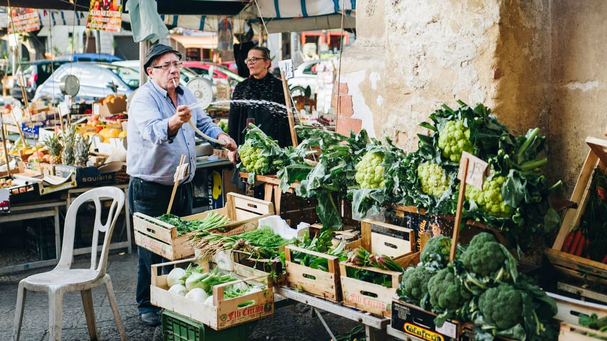 A male vendor wearing a flat cap and glasses waters fresh vegetables at a colorful outdoor market stall in Palermo, Sicily, while a woman stands in the background surrounded by crates of broccoli, cauliflower, and green onions.