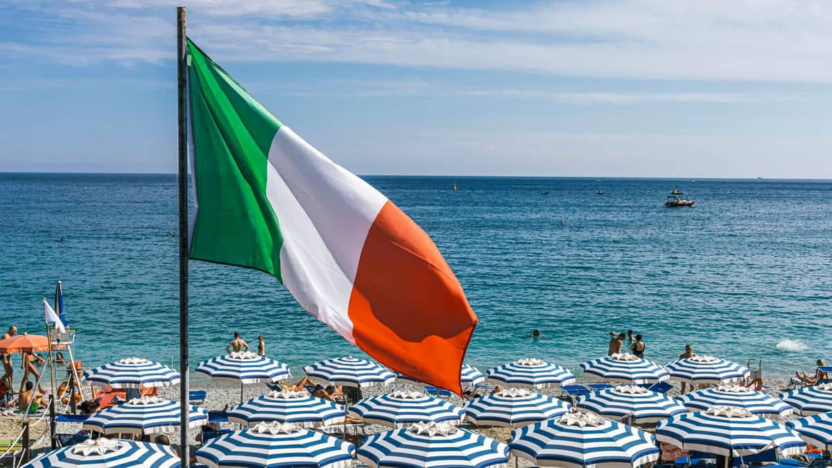 The Italian tricolor flag flies prominently over a busy Mediterranean beach lined with rows of blue-and-white striped umbrellas and sunbathers, with the calm turquoise sea and a boat visible in the background.