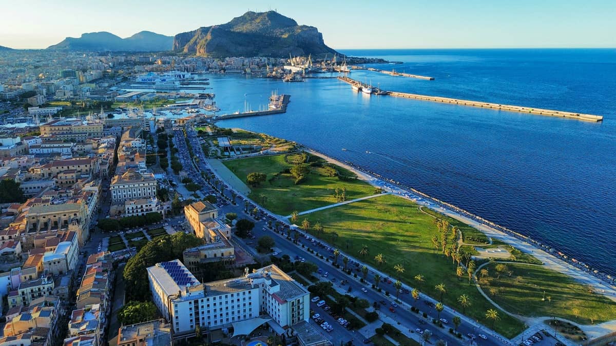 Aerial view of Palermo, Sicily at sunrise, showing the city's historic waterfront promenade, lush green coastal park, and busy harbor with Monte Pellegrino rising dramatically in the background against a clear blue sky.