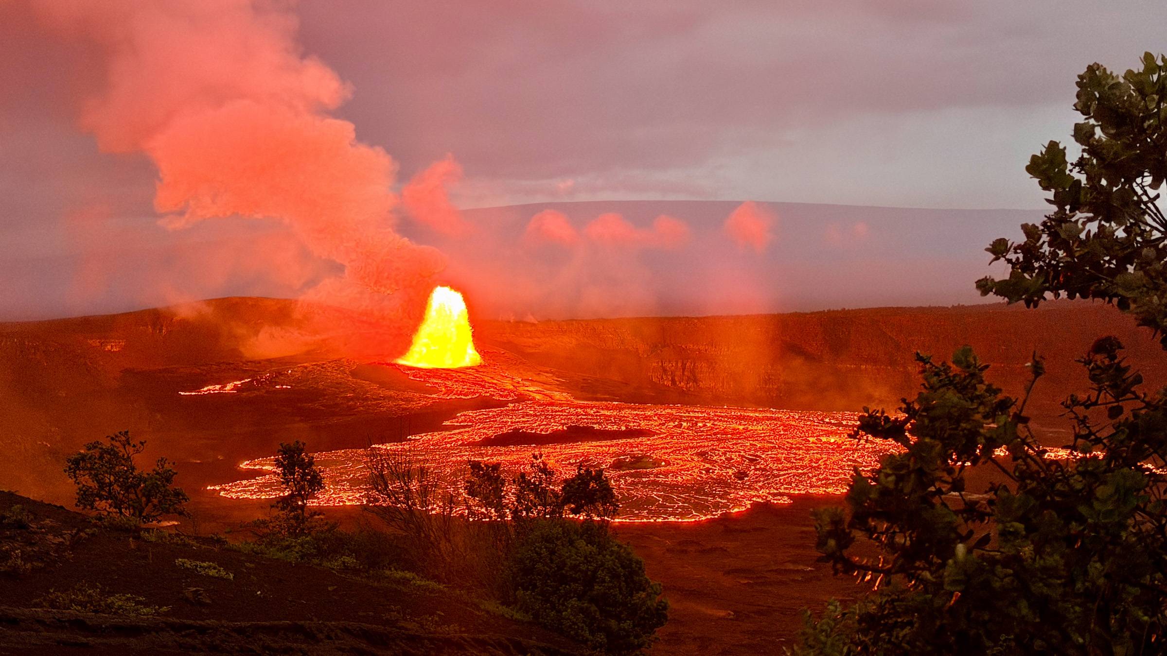 A dramatic wide-angle view of K?lauea volcano erupting at dusk, with a bright lava fountain shooting upward from the crater floor and a vast glowing lava lake spreading across the caldera, illuminating billowing volcanic plumes in fiery orange and red hue