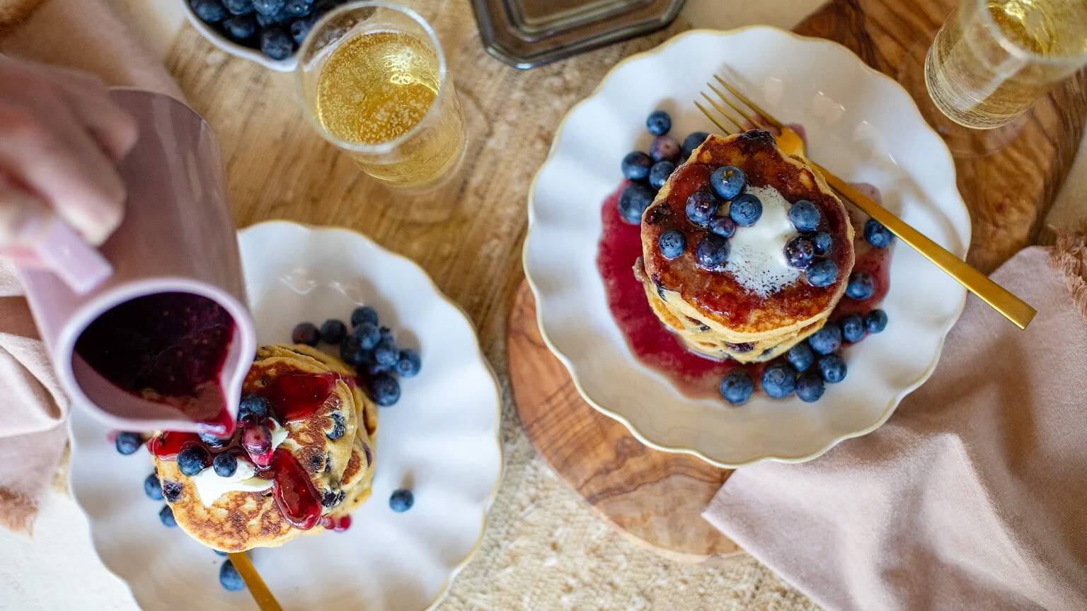 Overhead of two scalloped white plates stacked with blueberry pancakes topped with fresh blueberries and butter, as a hand pours a deep red blueberry compote over one stack, accompanied by glasses of sparkling wine on a wooden board.