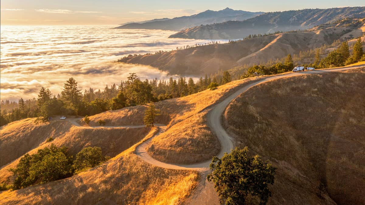 An aerial view of a winding dirt road snaking across sun-drenched golden hills above a dramatic sea of clouds in Big Sur, California, with forested ridges and distant mountain peaks bathed in warm early morning light.