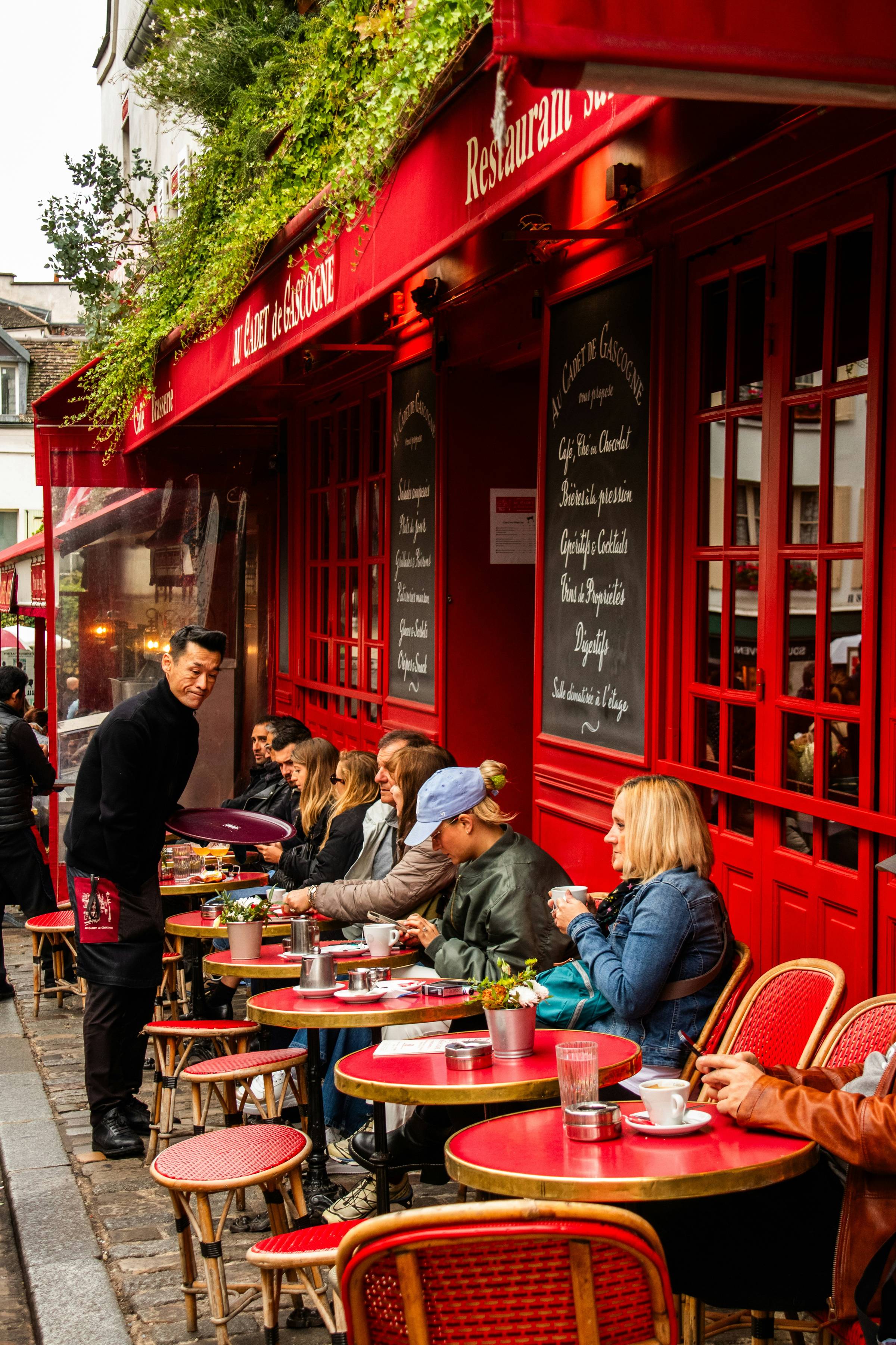 Patrons enjoy coffee and drinks at the outdoor terrace of Au Cadet de Gascogne, a classic Parisian café with a bold red facade in Montmartre, Paris, as a waiter carries a tray past the busy tables.