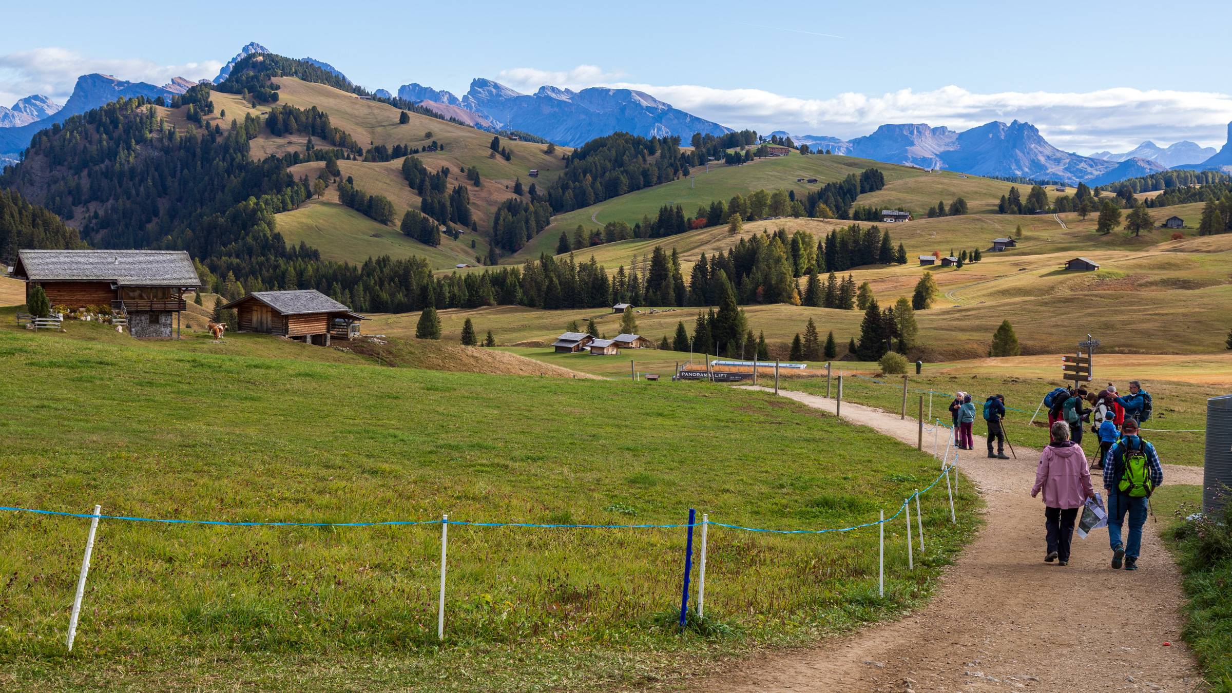 Two hikers walk along a dirt trail across the vast alpine meadows of Alpe di Siusi in South Tyrol, Italy, with rolling green hills, scattered mountain chalets, and the dramatic Dolomite peaks visible in the background under a clear blue autumn sky.