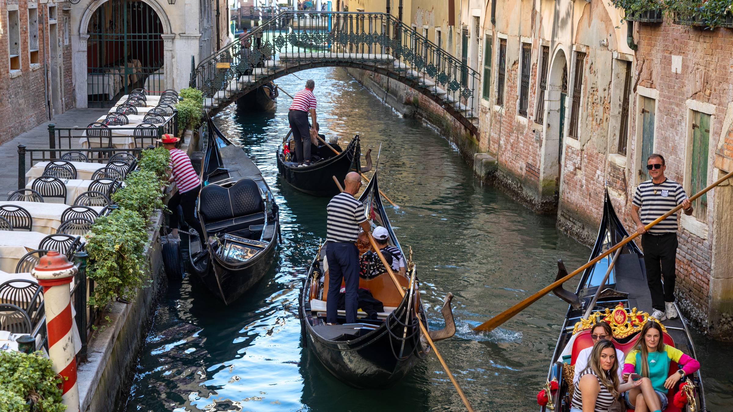 Multiple gondoliers in traditional striped shirts navigate black gondolas through a narrow Venetian canal lined with historic brick buildings, as female tourists enjoy a richly decorated gondola ride in Venice, Italy. An ornate iron footbridge arches over
