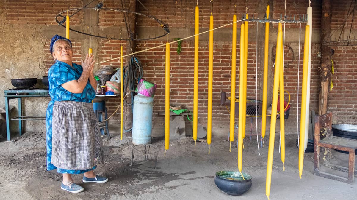 An elderly woman in a blue floral blouse and apron hand-dips long yellow beeswax candles suspended from a circular metal frame in a rustic brick workshop in Oaxaca, Mexico. A clay pot of melted yellow wax sits on the ground below the hanging candles.