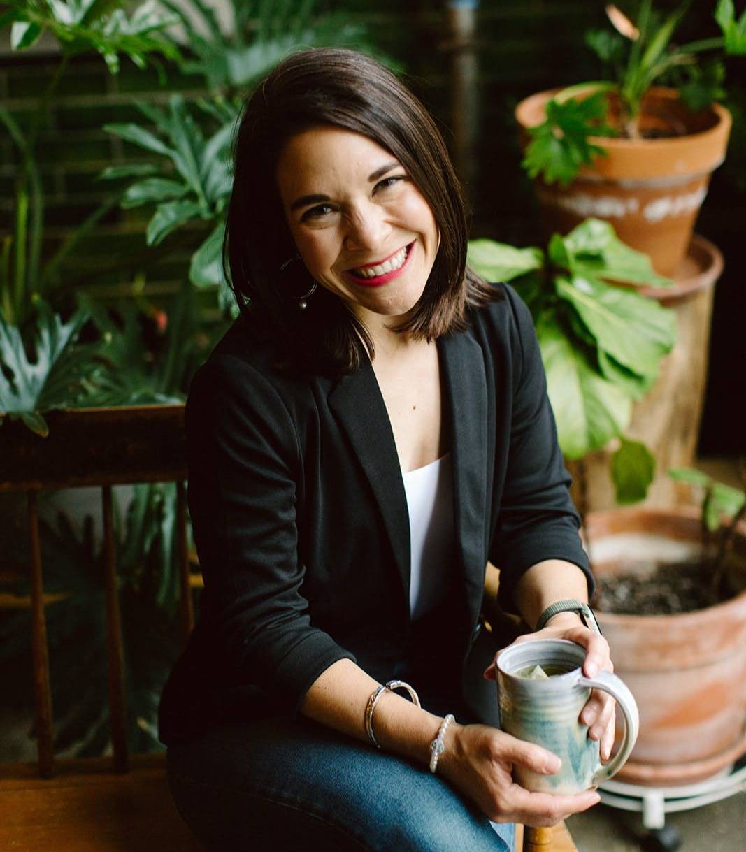 Kristina smiling and holding tea in front of houseplants. 