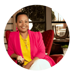 A person is sitting comfortably in a red armchair indoors. They are wearing a bright pink blazer over a yellow blouse and white pants, smiling warmly at the camera. The background shows a window with a view of trees and buildings outside, and there is a small plant on a table beside them.