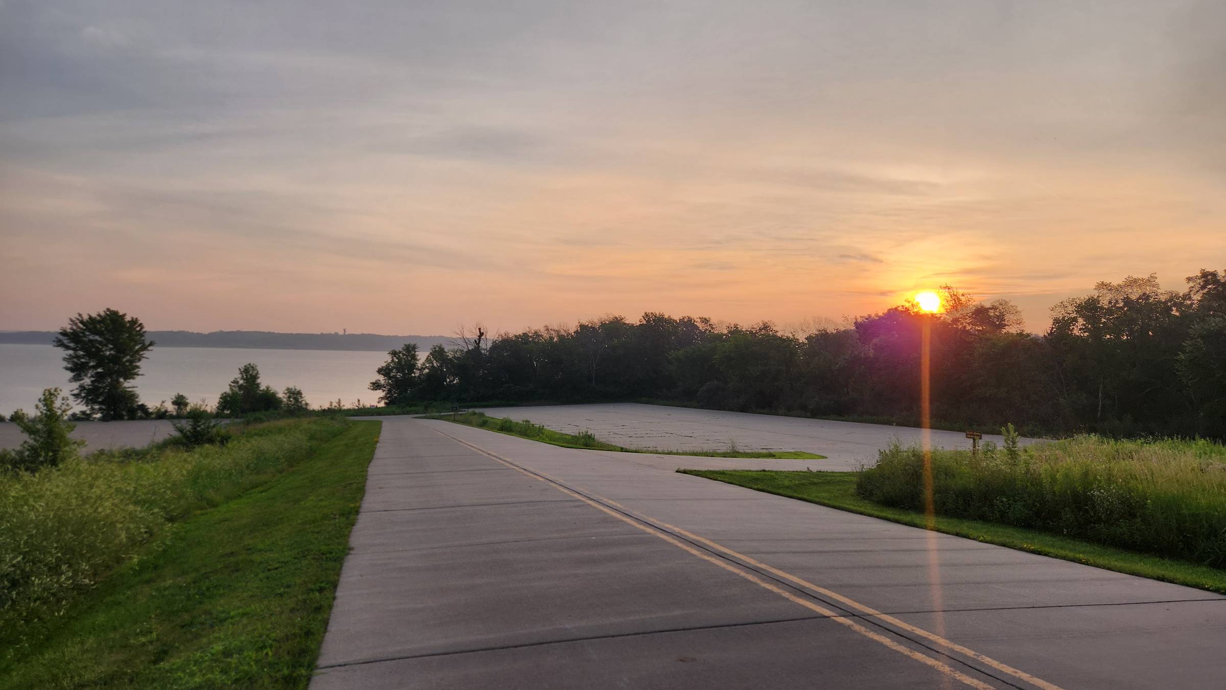 Peaceful country road at sunrise with golden light streaming through trees beside a calm lake