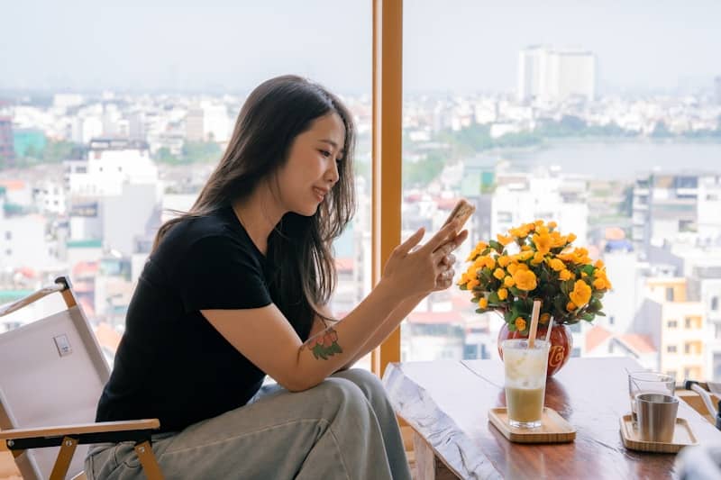 Young woman uses phone by window overlooking cityscape.