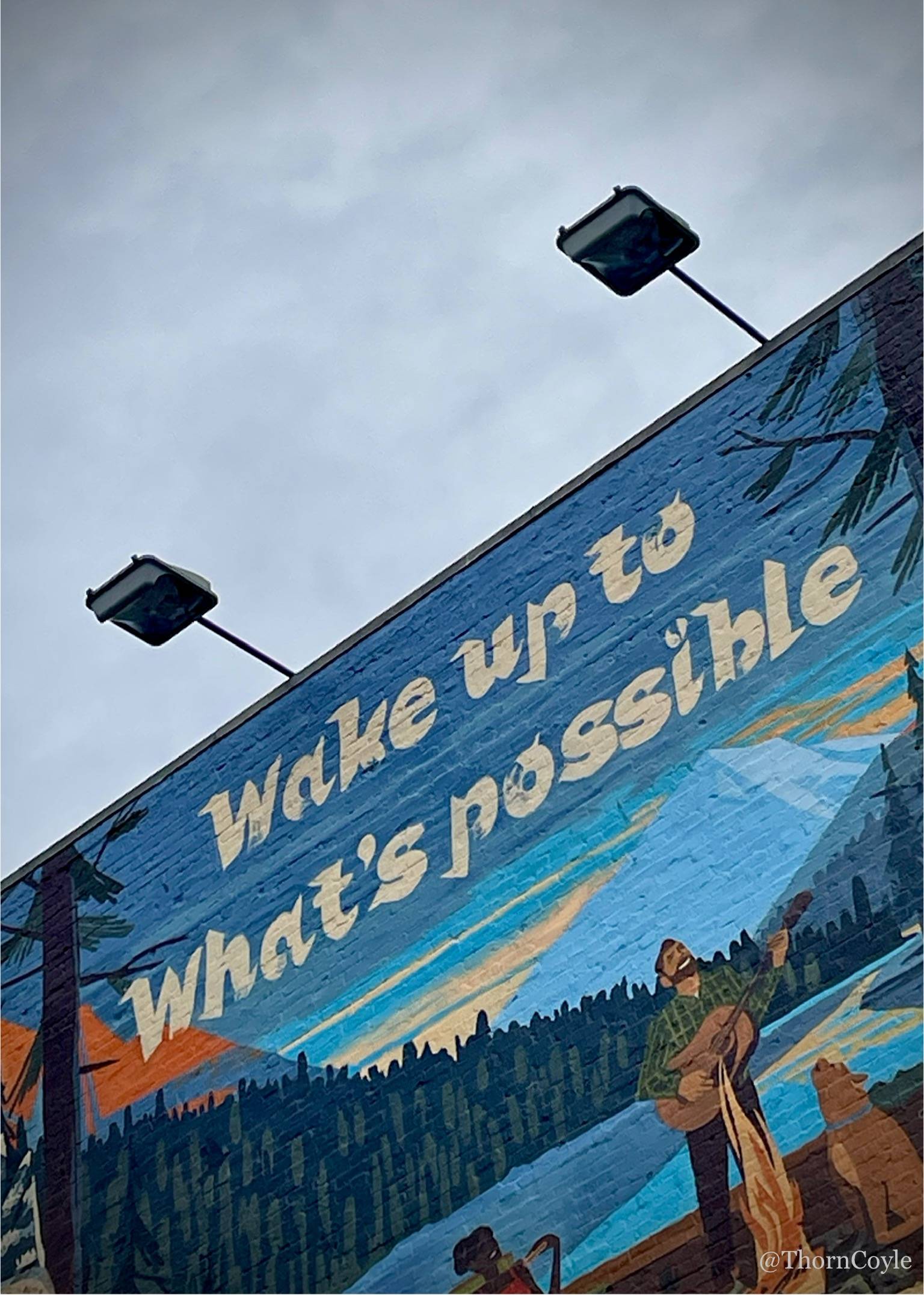 a wall mural under gray sky. Wake up to what's possible. a painted campfire and lake scene.