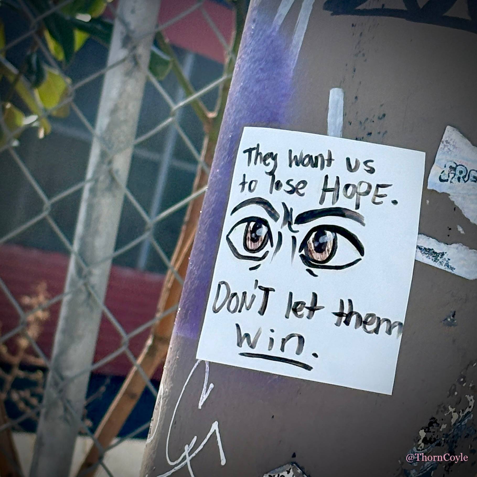 Hand drawn sticker on a metal utility pole near a chain link fence. Image of eyes. Text reads They want us to lose hope. Don’t let them win.