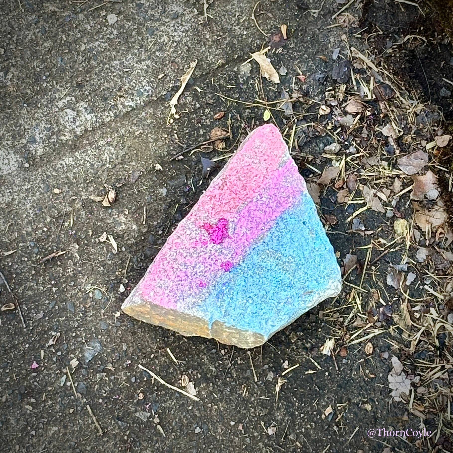 Photo: dirty sidewalk. A broken brick painted with the pale pink and blue stripes of the Trans Pride flag