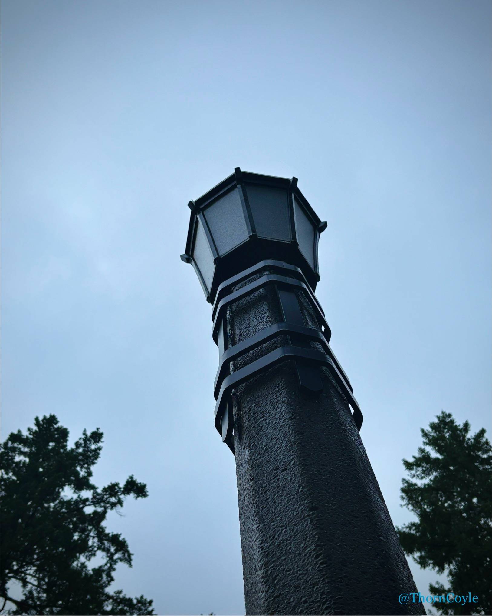 Looking up at an old fashioned street lamp, unlit beneath grey sky.