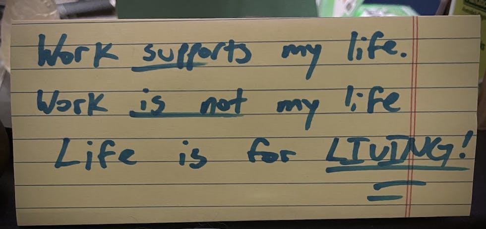 A photo of a folded piece of notebook paper with text written in blue marker. The text reads: "Work supports my life. Work is not my life. Life is for LIVING!":
