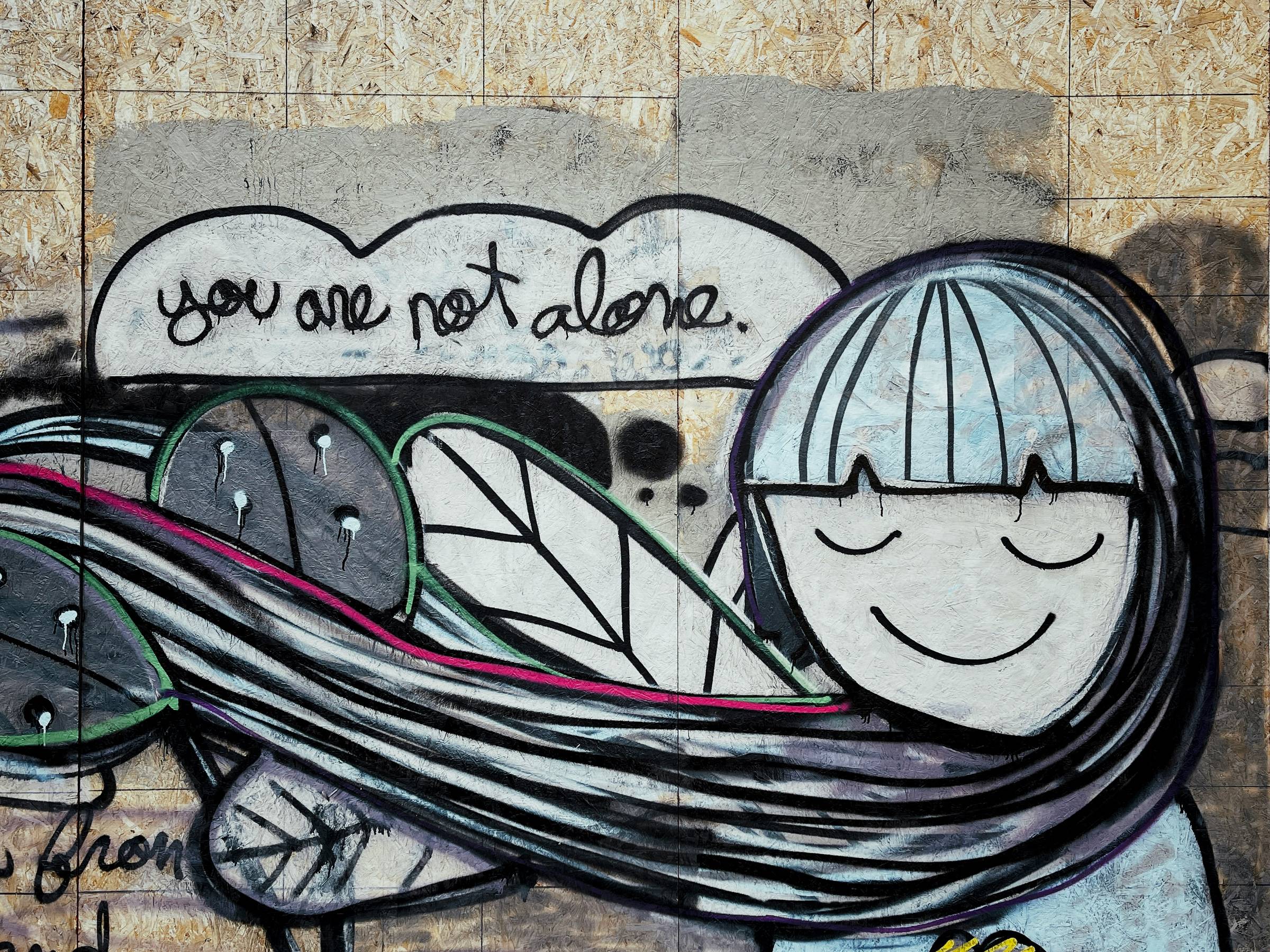 graffiti of young person with long hair and leafy arms holding clouds that say you are not alone