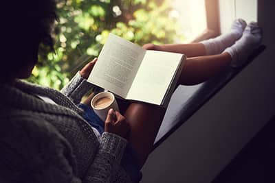 A woman sits near a window, engrossed in a book, holding a mug of tea or coffee.