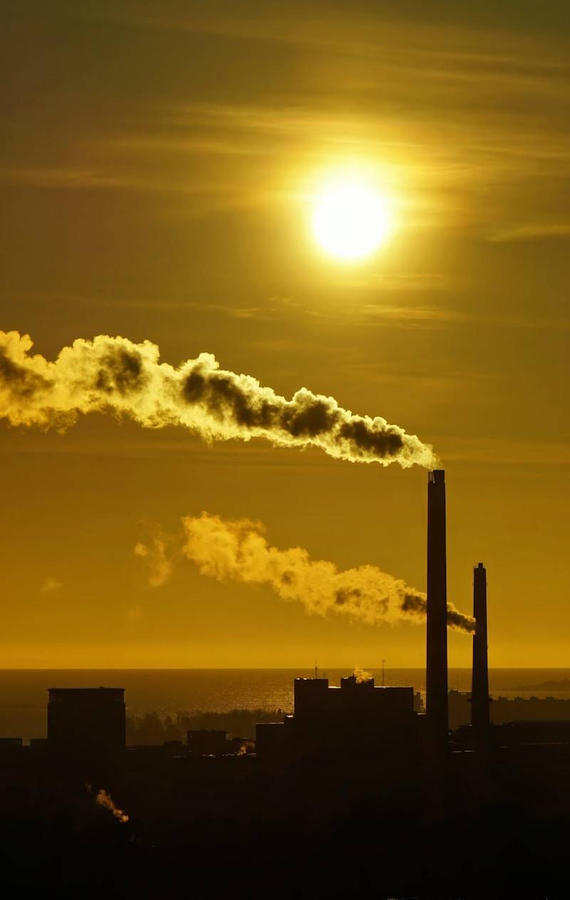 The sun is setting behind a factory with smoke coming out of it