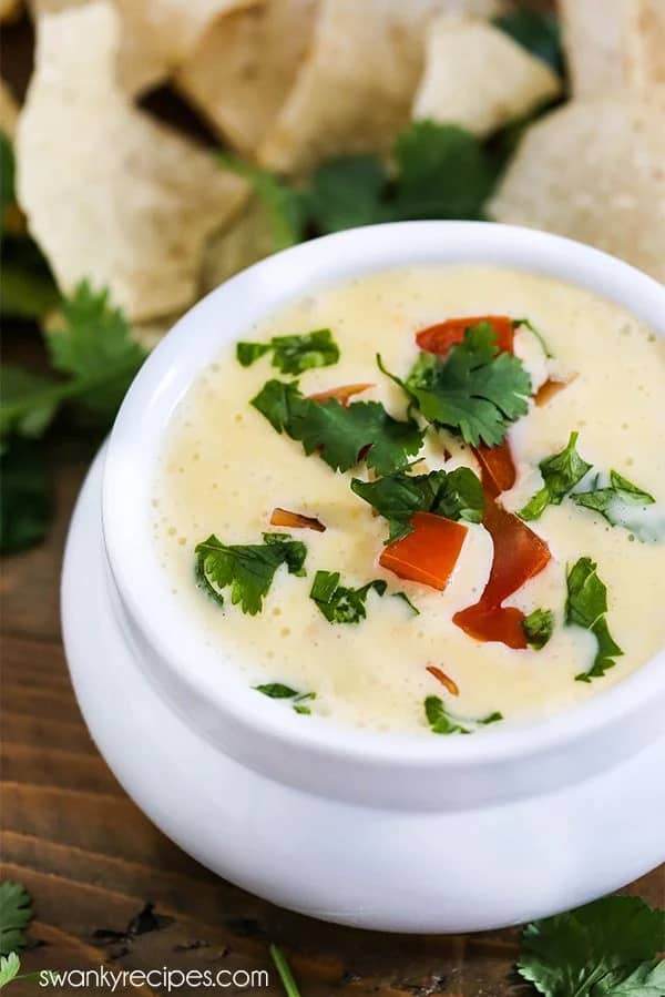 Bowl of creamy Mexican queso dip topped with cilantro and red pepper, surrounded by tortilla chips.