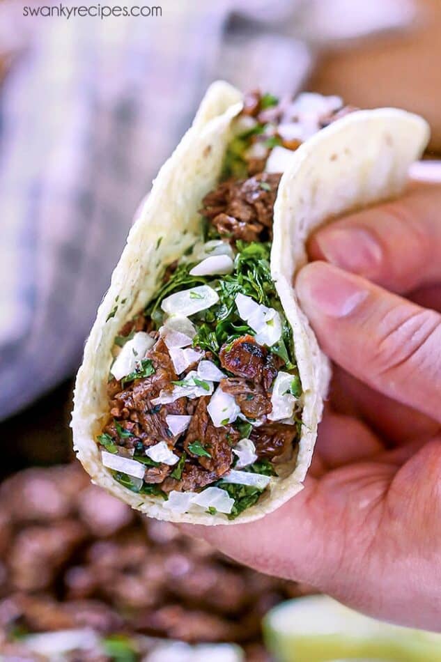 Hand holding a carne asada taco with grilled beef, cilantro, and diced onions in a soft tortilla.