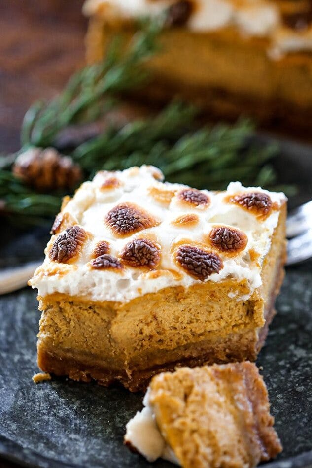 A pumpkin pie bar on a plate with a slice cut off. Pumpkin pie bars feature a pumpkin pie center with toasted marshmallows on top.
