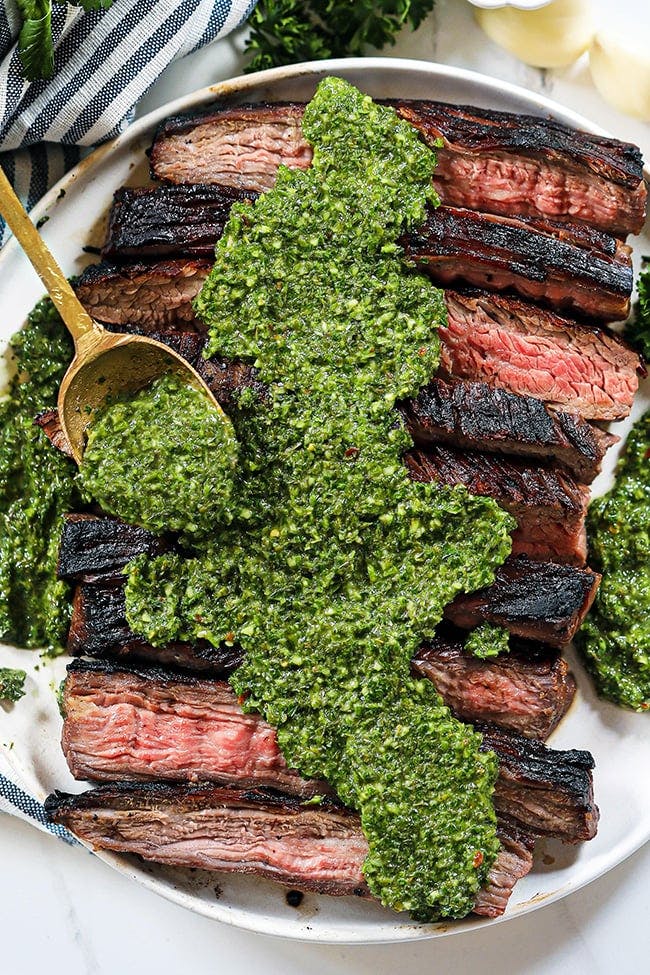 Flank steak cut into long pieces showed on a plate with a heaping spoonful of chimichurri sauce on top of the beef meat.