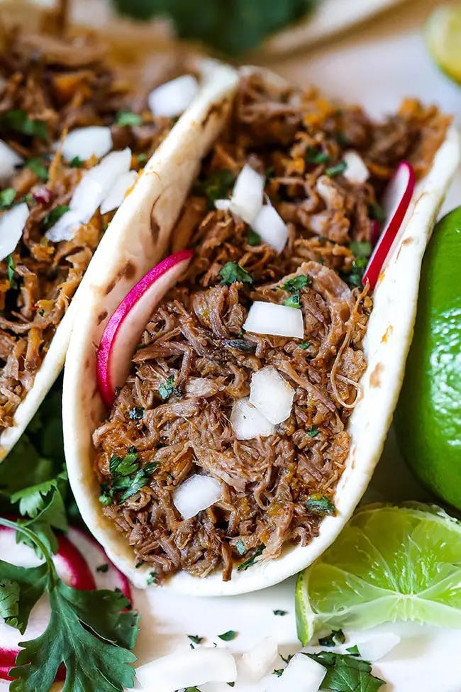 A flour tortilla loaded with shredded beef barbacoa, topped with diced white onions, thin slices of radish, and fresh cilantro leaves.