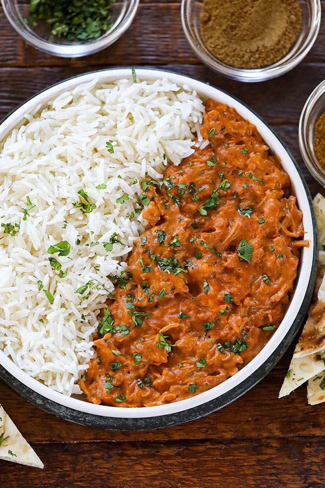 A flavorful bowl of Chicken Tikka Masala, garnished with spices and served on a wooden board. The dish is complemented with perfectly cooked rice, and surrounded by an array of spices used in its preparation.
