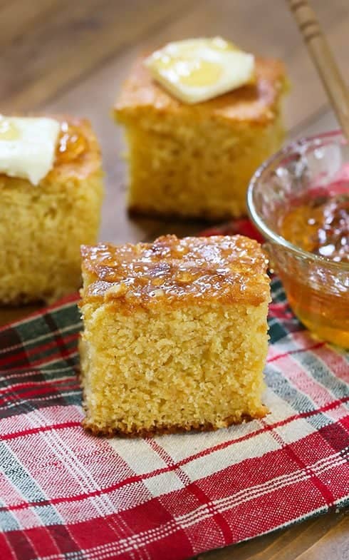 Square pieces of sweet buttermilk cornbread on a red plaid cloth, topped with butter and served with honey.