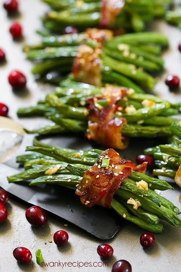 A tantalizing image of green beans bundled together with bacon, seasoned with garlic and pepper flakes, and garnished with cranberries. The dish is artfully arranged on a wooden cutting board, ready to be served.