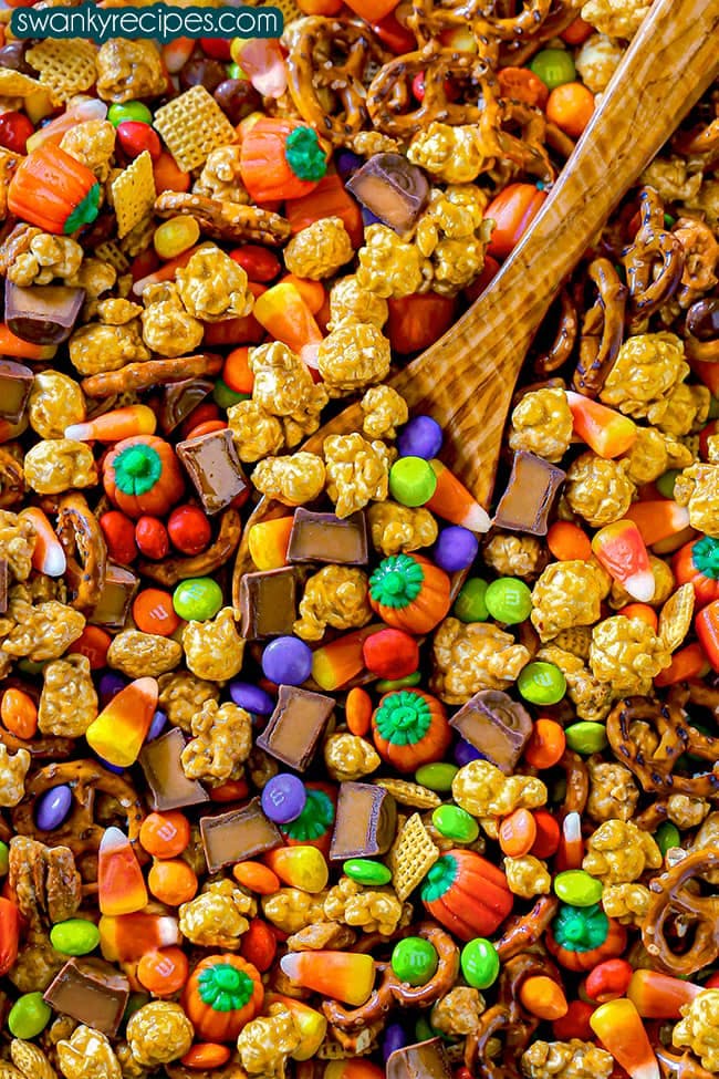 A white bowl filled with mostly caramel corn, Red, purple, orange, and brown M&Ms sprinkled throughout the bowl with orange and green pumpkins, classic candy corn, and yellow chex mix.