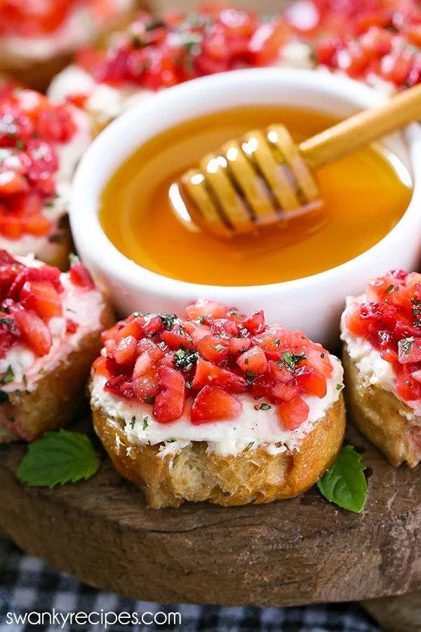 A plate of Strawberry Bruschetta with toasted slices of bread that is topped with a layer of cream cheese,  freshly diced strawberries, chopped mint, and a drizzle of honey.