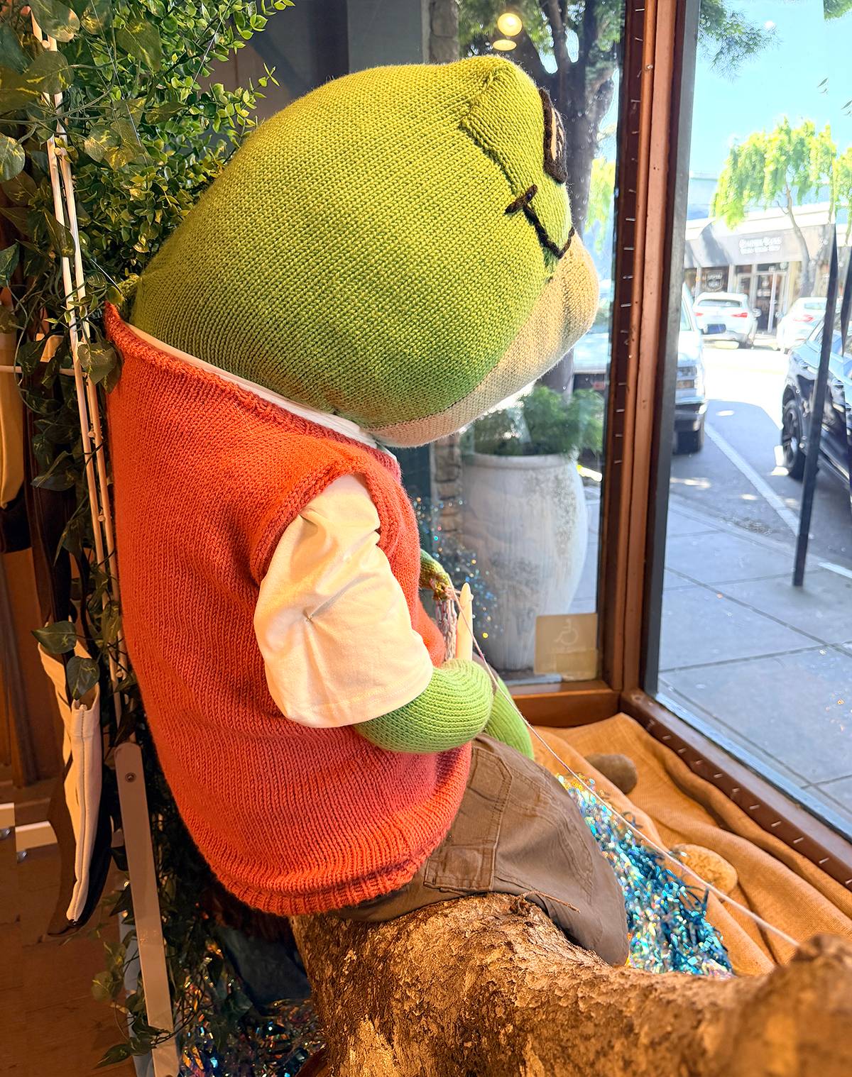 side view of knitted Karl the Frog wearing vest and shorts looking out yarn shop window at Fengari in Half Moon Bay.