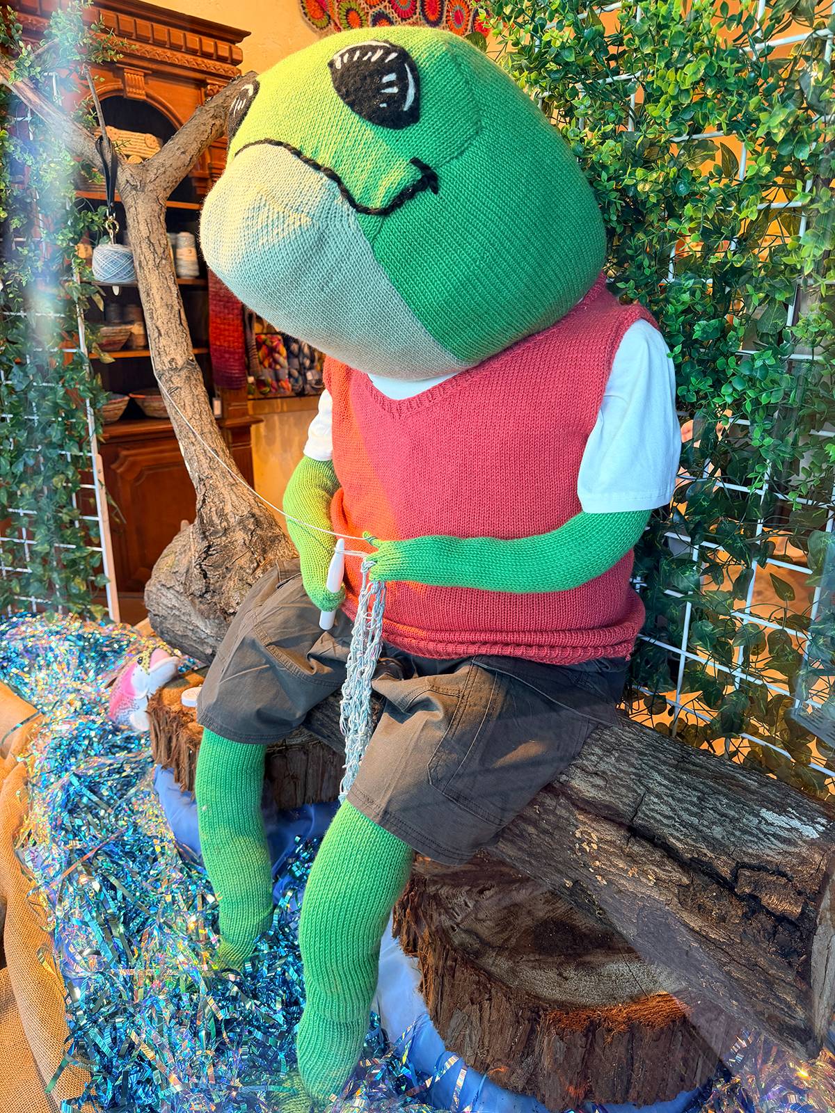 large knitted Karl the Frog mascot sitting on log knitting in Fengari yarn shop window display.